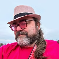 Une personne aux cheveux longs, barbu et portant des lunettes sourit, coiffée d'un chapeau rose et vêtue d'une chemise rouge. Le visage calme, elle se trouve à l'extérieur sous un ciel nuageux. Une personne aux cheveux longs, barbu et portant des lunettes sourit, coiffée d'un chapeau rose et vêtue d'une chemise rouge. Le visage calme, elle se trouve à l'extérieur sous un ciel nuageux.