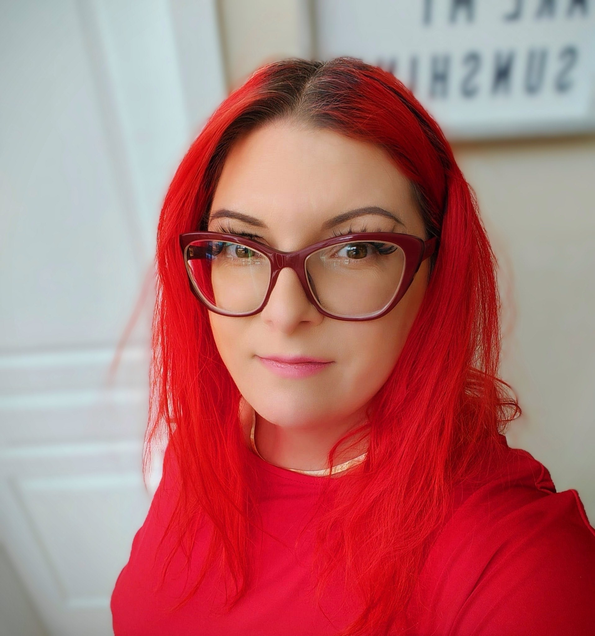 Close-up of a person with bright red hair and large glasses, smiling. They wear a red top and stand in front of a softly blurred indoor background with white walls Close-up of a person with bright red hair and large glasses, smiling. They wear a red top and stand in front of a softly blurred indoor background with white walls