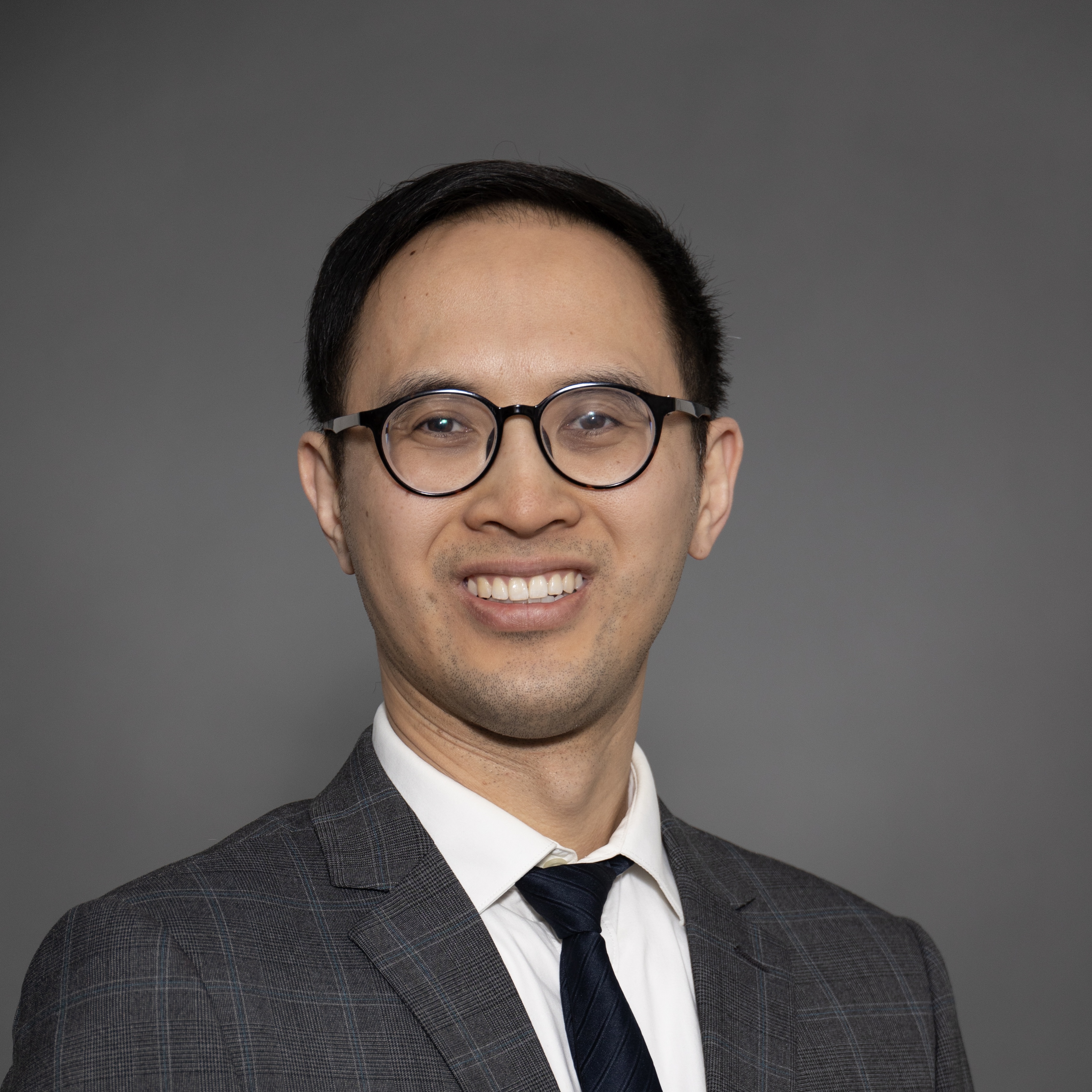 Professional headshot of a smiling man with short hair, glasses, and a checkered suit jacket over a white shirt and dark tie, in front of a grey background. Professional headshot of a smiling man with short hair, glasses, and a checkered suit jacket over a white shirt and dark tie, in front of a grey background.