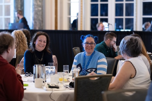 Four people sit at a round table, smiling and engaged in conversation in a conference room. One participant wears a blue cat sweater and lanyard. Glasses of water and notepads are on the table. Four people sit at a round table, smiling and engaged in conversation in a conference room. One participant wears a blue cat sweater and lanyard. Glasses of water and notepads are on the table.