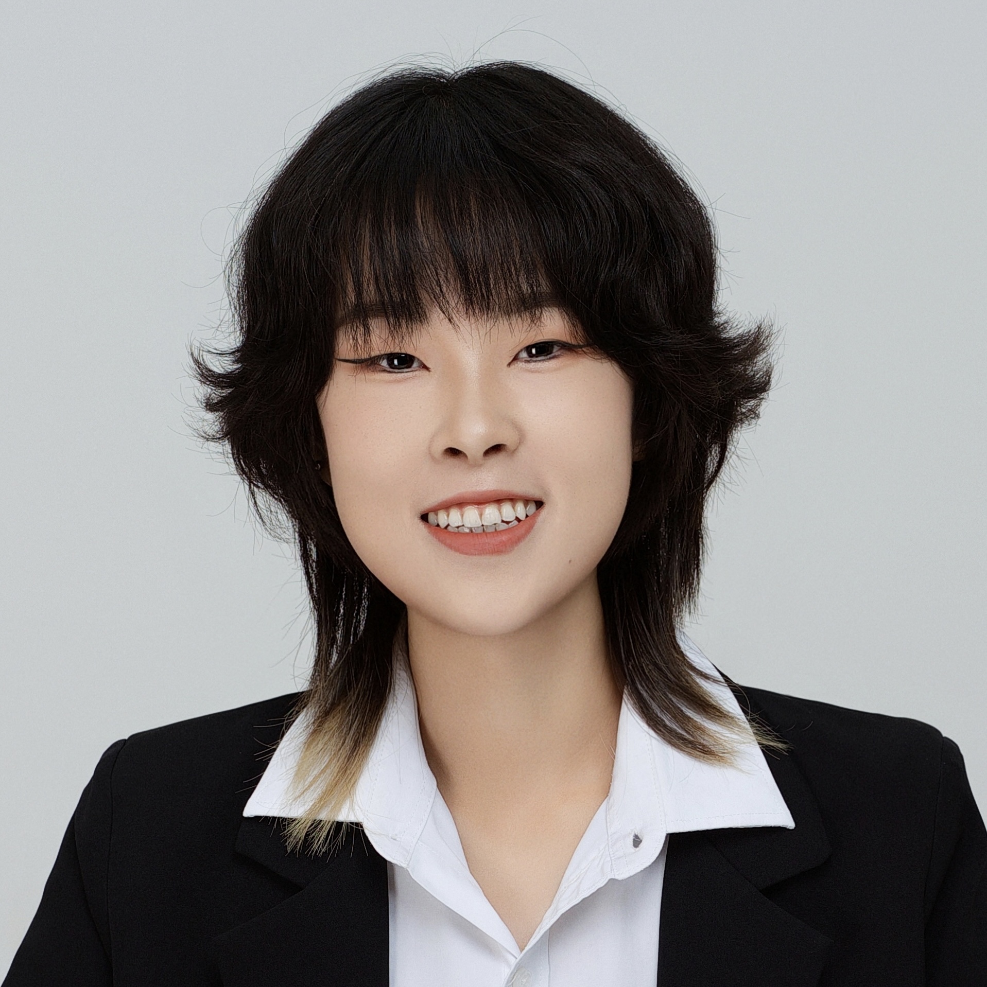 Portrait professionnel d'une personne aux cheveux courts foncés dégradés avec une mèche blonde, souriante, vêtue d'une chemise blanche et d'un blazer noir, sur fond clair. Portrait professionnel d'une personne aux cheveux courts foncés dégradés avec une mèche blonde, souriante, vêtue d'une chemise blanche et d'un blazer noir, sur fond clair.