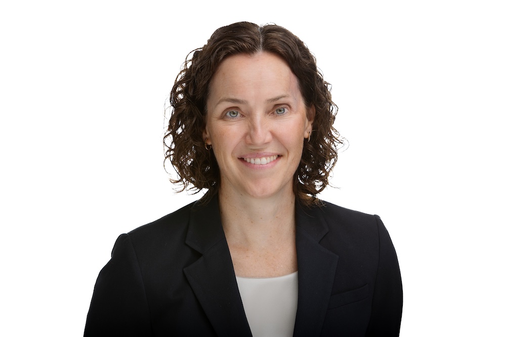 Professional headshot of a smiling woman with curly brown hair, wearing a black blazer and light blouse, on a white background. Professional headshot of a smiling woman with curly brown hair, wearing a black blazer and light blouse, on a white background.