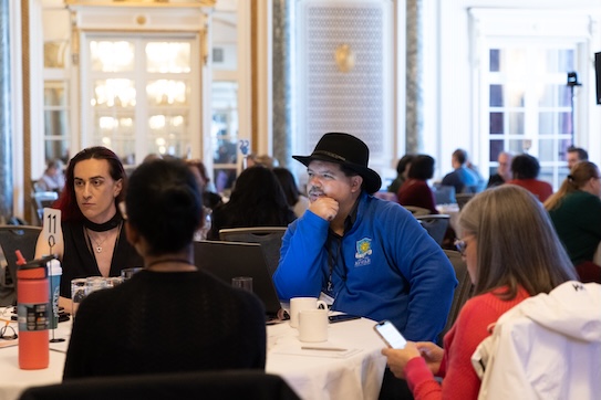 A group of people sitting at a table in a bright, elegant room. One person in a black hat and blue hoodie listens thoughtfully. Others are using laptops or phones. A group of people sitting at a table in a bright, elegant room. One person in a black hat and blue hoodie listens thoughtfully. Others are using laptops or phones.