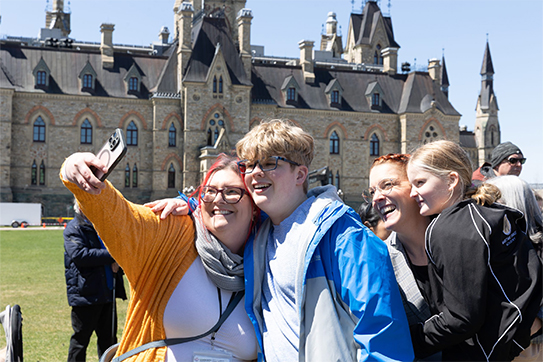 A group of four people smile while taking a selfie in front of the Parliament buildings in Ottawa. They appear joyful, standing close together on a sunny day during an autism community event. A group of four people smile while taking a selfie in front of the Parliament buildings in Ottawa. They appear joyful, standing close together on a sunny day during an autism community event.