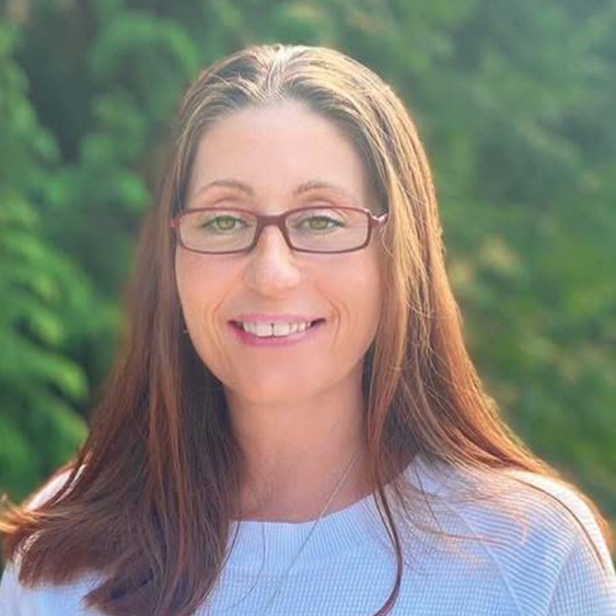 Photo professionnelle d'une femme souriante aux cheveux foncés et portant des lunettes, vêtue d'un blazer bleu et d'une chemise rayée, devant un fond blanc.