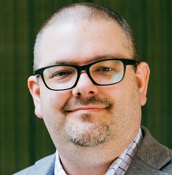 Photo en noir et blanc d'une femme aux longs cheveux ondulés et portant des lunettes, souriant doucement devant un fond blanc texturé.