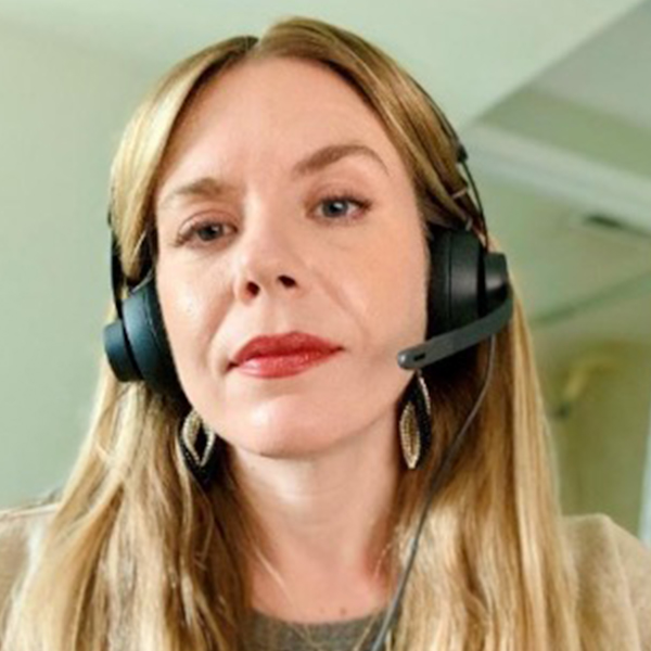A person with long hair, a beard, and glasses smiles while wearing a pink hat and red shirt. They have a calm expression and are outdoors under a cloudy sky.