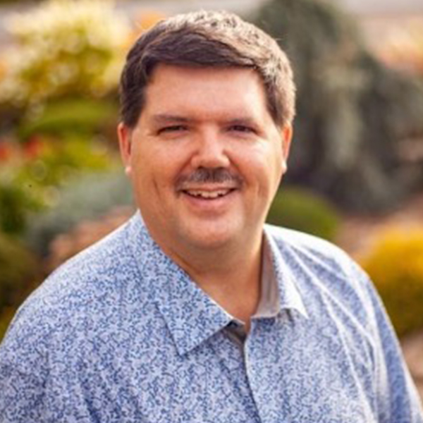 A person with long hair, a beard, and glasses smiles while wearing a pink hat and red shirt. They have a calm expression and are outdoors under a cloudy sky.