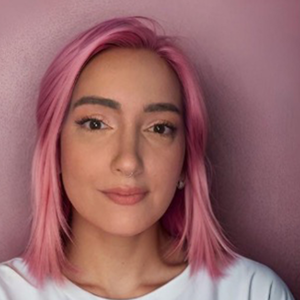 Professional headshot of a person with short layered dark hair and a streak of blonde, smiling, wearing a white shirt and black blazer, against a pale background.