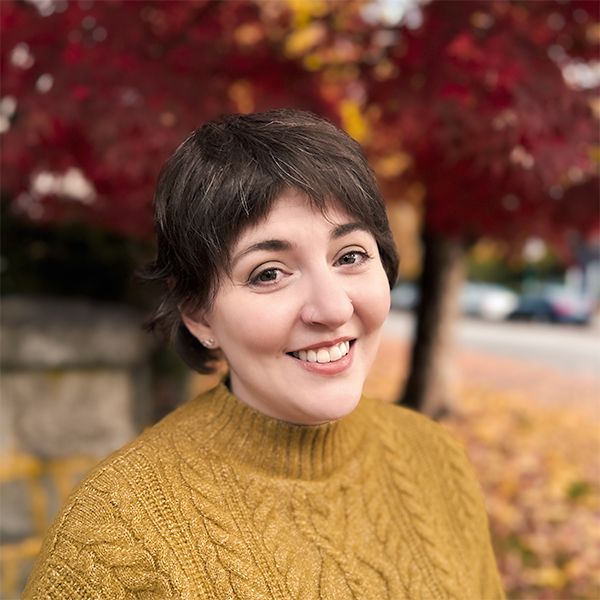 Professional headshot of a smiling woman with dark hair and glasses, wearing a blue blazer and striped shirt, in front of a white background.