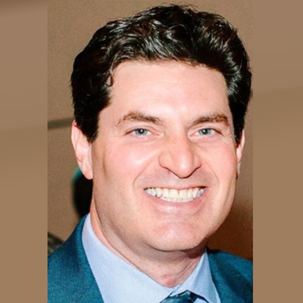 Professional headshot of a smiling man with short hair, glasses, and a checkered suit jacket over a white shirt and dark tie, in front of a grey background.