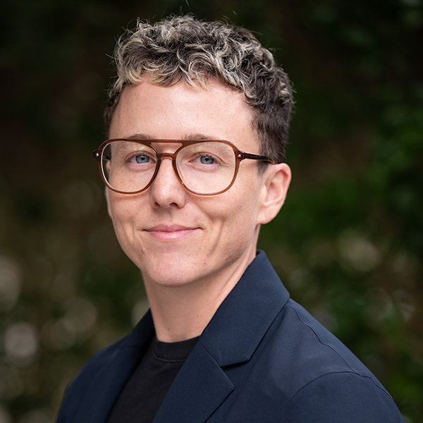 Close-up of a person with bright red hair and large glasses, smiling. They wear a red top and stand in front of a softly blurred indoor background with white walls