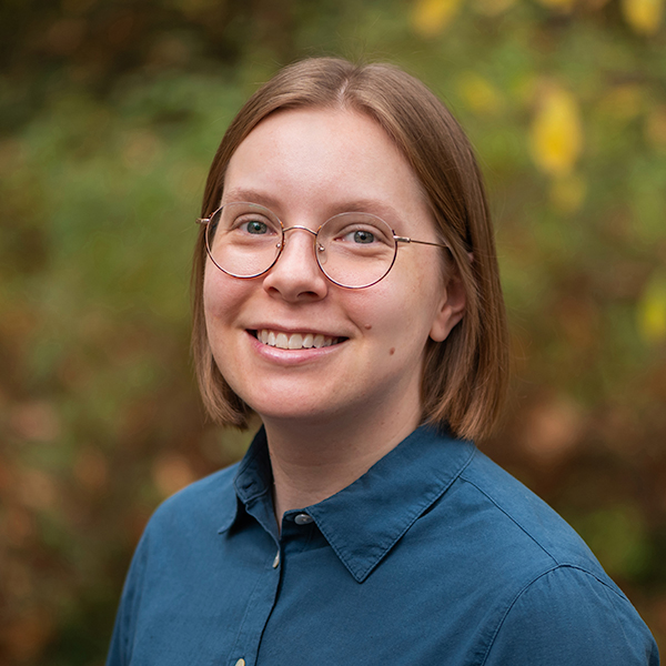 Black and white headshot of a woman with long wavy hair and glasses, smiling gently standing in front of a textured white background.