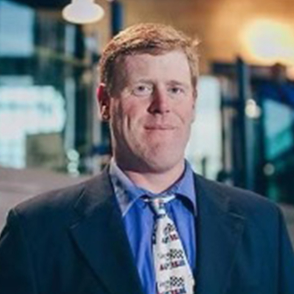 Professional headshot of a person with short layered dark hair and a streak of blonde, smiling, wearing a white shirt and black blazer, against a pale background.