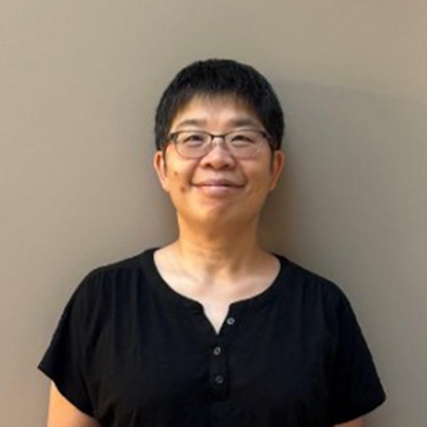 A person with long hair, a beard, and glasses smiles while wearing a pink hat and red shirt. They have a calm expression and are outdoors under a cloudy sky.
