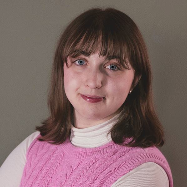 Professional headshot of a smiling woman with dark hair and glasses, wearing a blue blazer and striped shirt, in front of a white background.