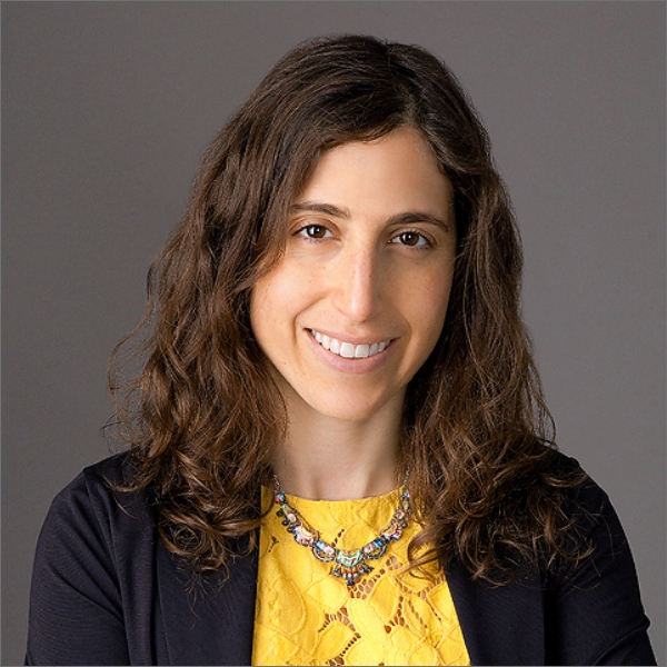 A person with long hair, a beard, and glasses smiles while wearing a pink hat and red shirt. They have a calm expression and are outdoors under a cloudy sky.