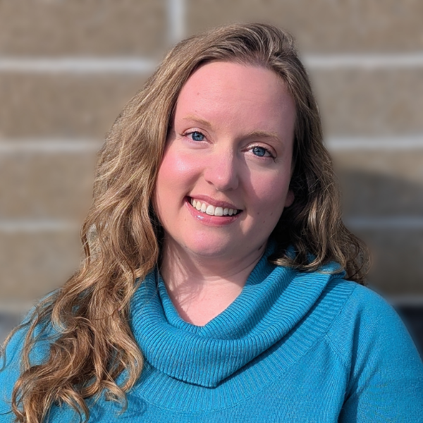 A person with long hair, a beard, and glasses smiles while wearing a pink hat and red shirt. They have a calm expression and are outdoors under a cloudy sky.