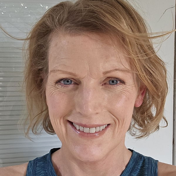 A person with long hair, a beard, and glasses smiles while wearing a pink hat and red shirt. They have a calm expression and are outdoors under a cloudy sky.