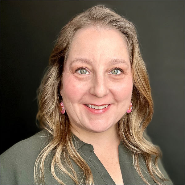 Professional headshot of a person with short layered dark hair and a streak of blonde, smiling, wearing a white shirt and black blazer, against a pale background.
