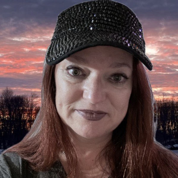 A person with long hair, a beard, and glasses smiles while wearing a pink hat and red shirt. They have a calm expression and are outdoors under a cloudy sky.