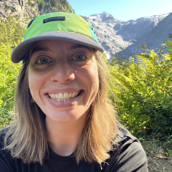 Professional headshot of a person with short layered dark hair and a streak of blonde, smiling, wearing a white shirt and black blazer, against a pale background.