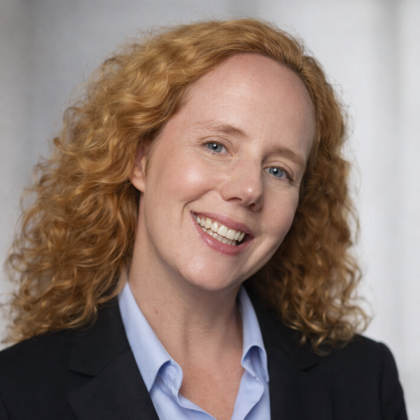 A person with long hair, a beard, and glasses smiles while wearing a pink hat and red shirt. They have a calm expression and are outdoors under a cloudy sky.