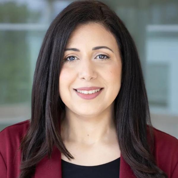 A person with long hair, a beard, and glasses smiles while wearing a pink hat and red shirt. They have a calm expression and are outdoors under a cloudy sky.